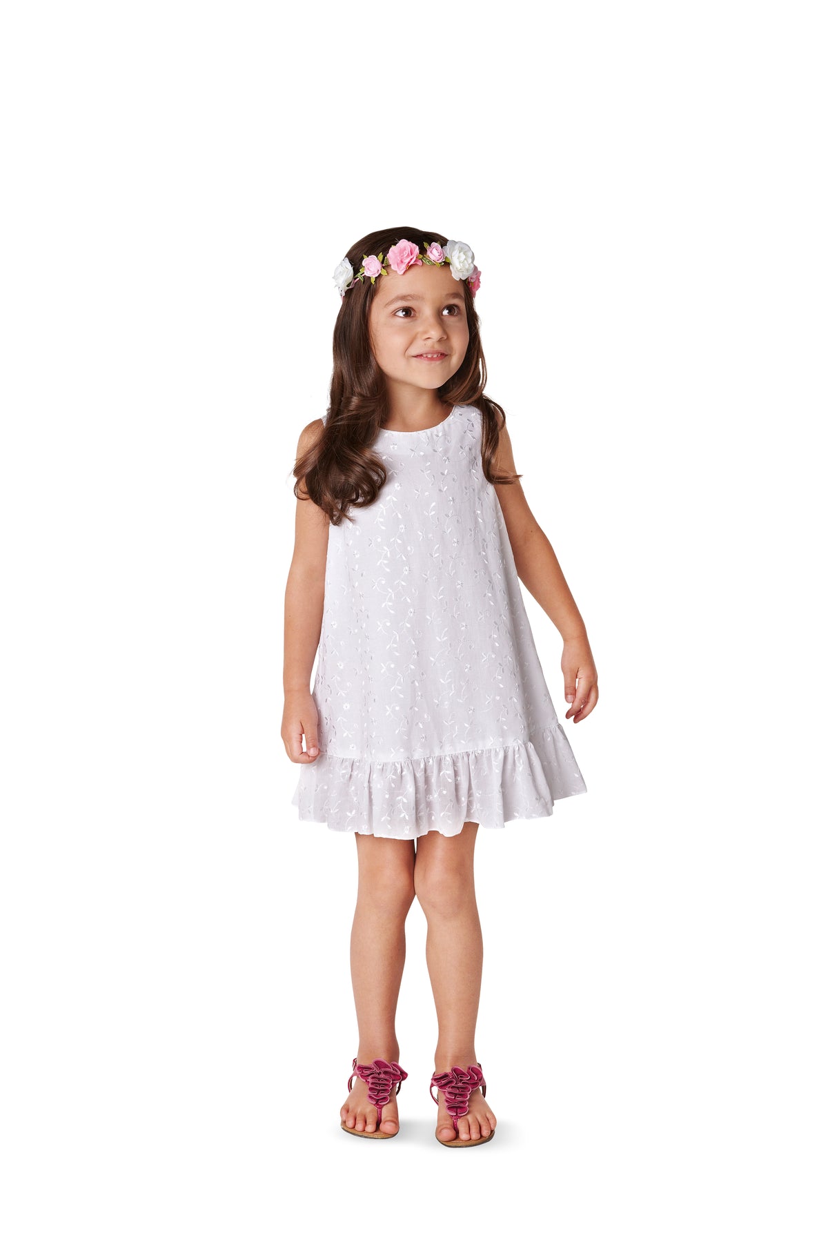 Petite fille en robe blanche à volants, avec diadème floral et sandales roses, sourire léger.