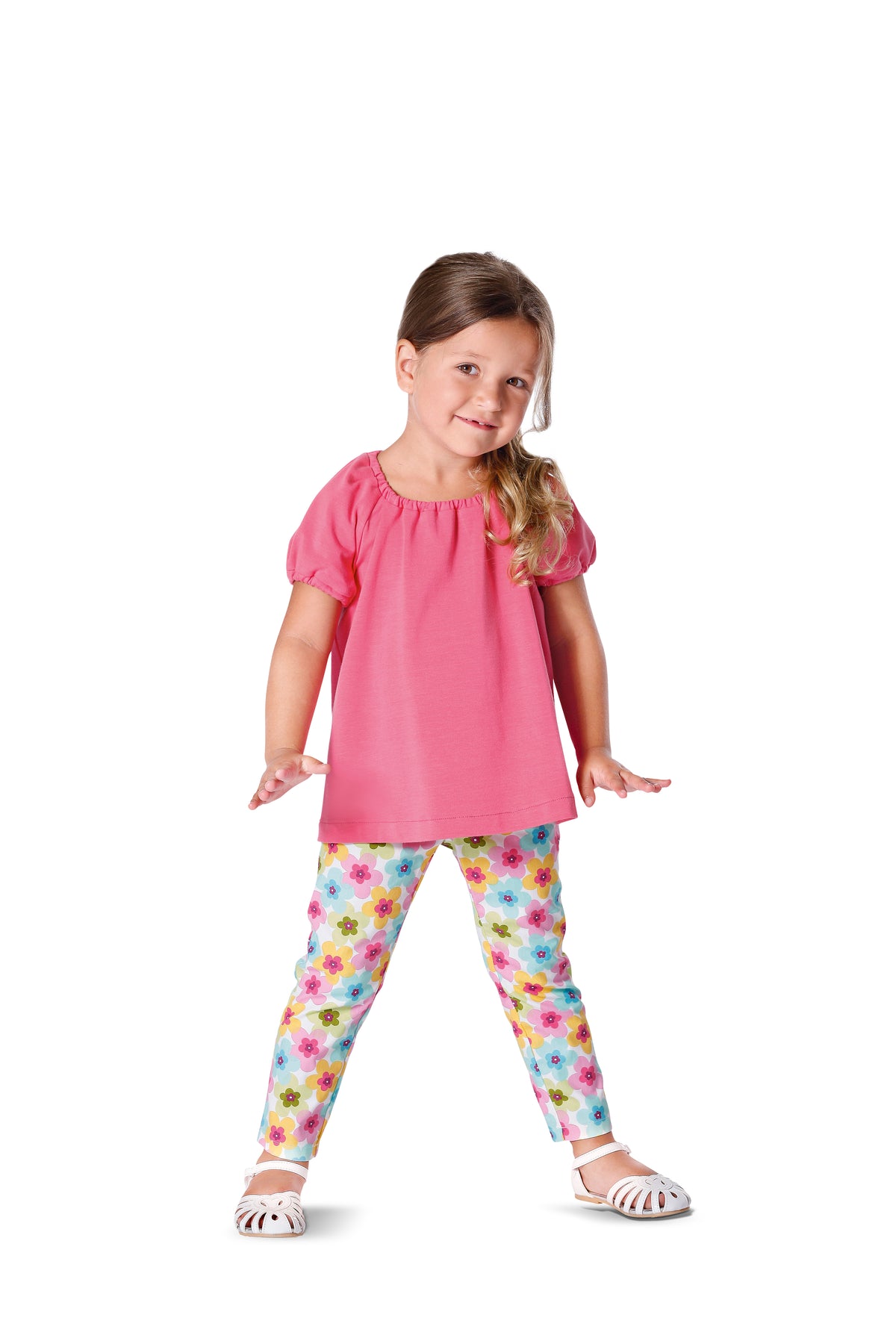Petite fille en t-shirt rose et leggings fleuris, pieds en sandalettes blanches, posant joyeusement.