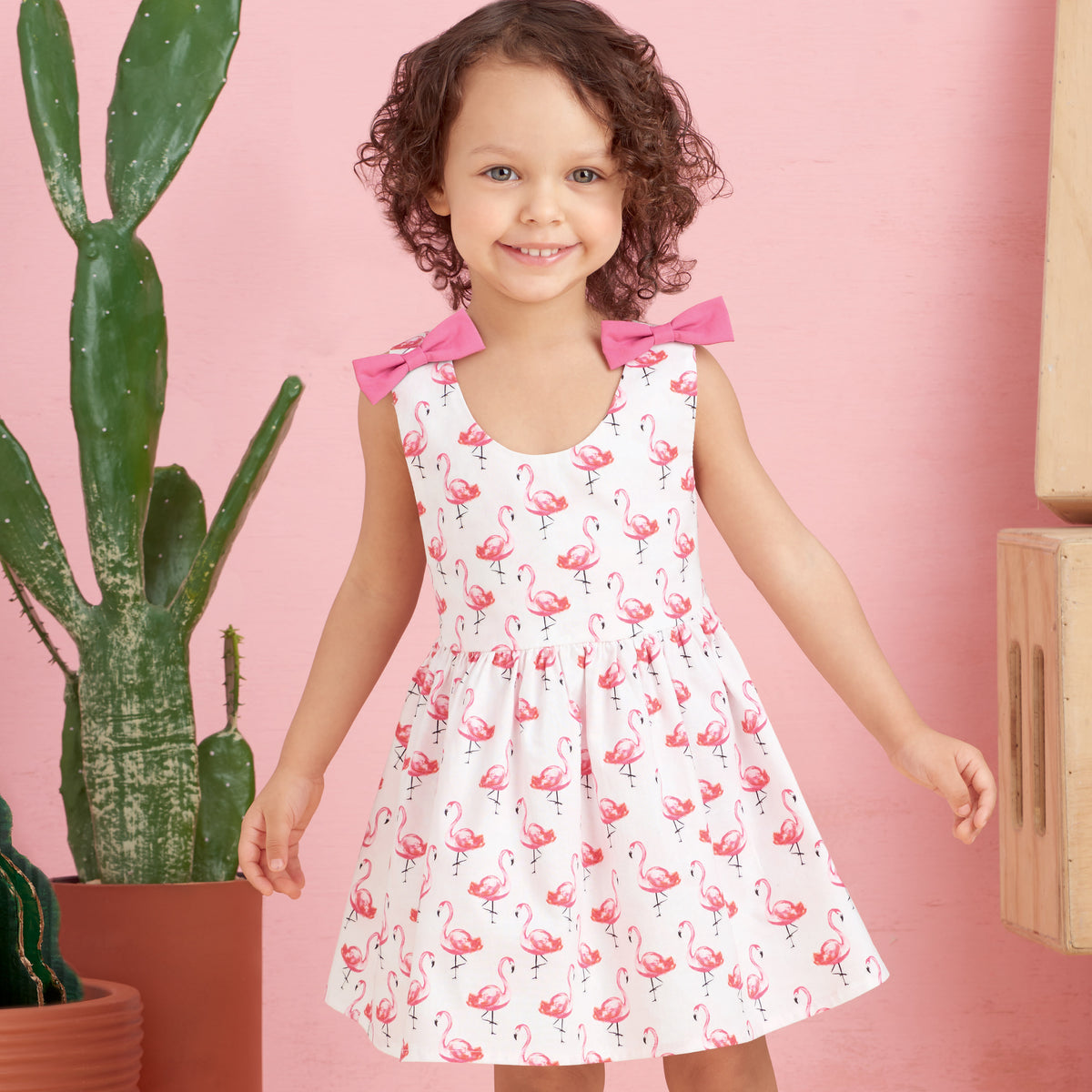 Jeune fille souriante portant une robe blanche à flamants roses, clips roses sur les épaules, debout près d’un mur rose avec des plantes cactus à gauche.
