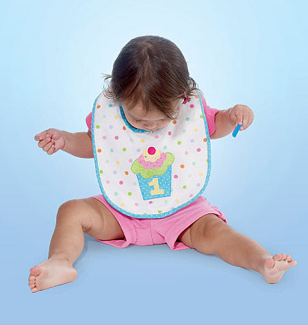 Bébé assise sur fond bleu, bavette blanche à pois colorés et motif gâteau numéro 1, vêtements roses, tient une cuillère bleue.