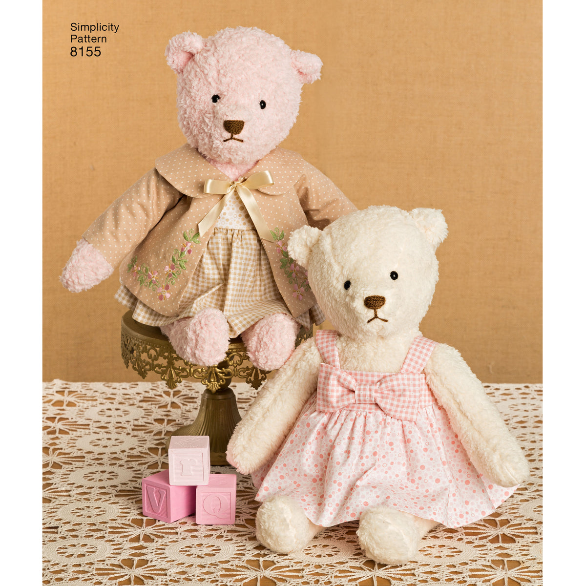 Deux doudous ours vintage en peluche près d’un cadre beige: l’un debout sur un socle et l’autre assis, habillés de robes pastel à motifs, avec blocs roses sur la nappe crochetée.